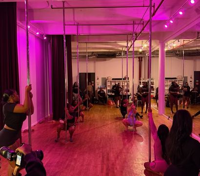 Energetic pole fitness class in a pink-lit studio — chrome poles, hardwood floor and mirrored wall with participants practicing moves while a photographer shoots.