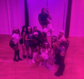 Group of women striking playful poses for a mirror selfie during a pole dance/fitness class in a dance studio bathed in pink-purple lighting