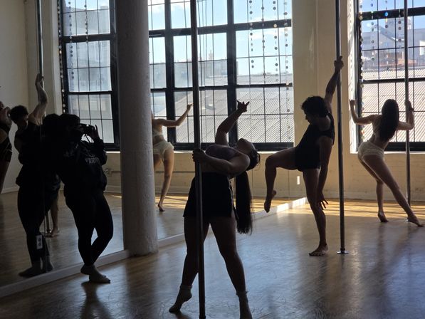 Sunlit loft pole fitness class with dancers striking dramatic poses around chrome poles by large industrial windows and mirrored wall, city rooftop view