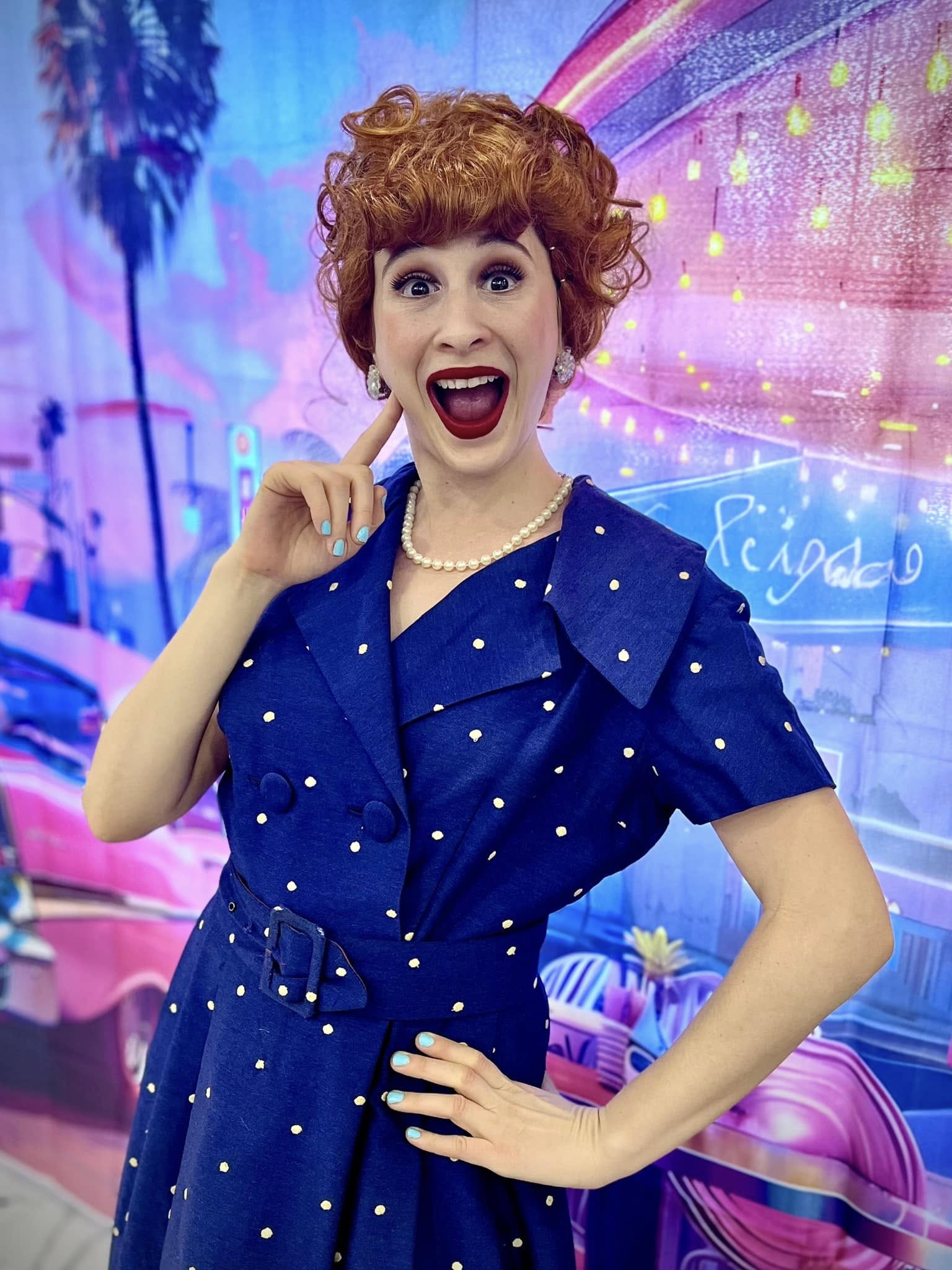 Excited redhead in a vintage 1950s navy polka-dot dress with pearl necklace and bright red lipstick, posing with hand on her cheek against a colorful neon retro diner backdrop with palm trees.