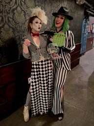 Two people in black-and-white carnival costumes pose in a dim hallway against damask wallpaper — one with white face paint, feathered headpiece and harlequin check skirt; the other with a green wig, fedora, striped suit holding a bingo card and a small bingo cage with colorful balls.