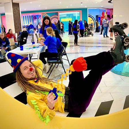 Colorful clown with polka-dot hat, red nose and roller skates lounging on a yellow crescent bench at a busy indoor shopping mall family event, holding a toy grabber with a red foam hand as volunteers and families gather at activity tables and vibrant storefronts in the background.
