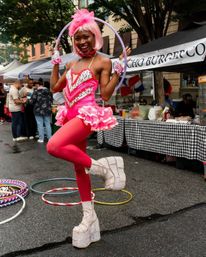 Performer in a bright pink wig and sequined tutu poses with a hula hoop at an outdoor street fair, red tights and glitter platform boots with vendor tents and extra hoops in the background.