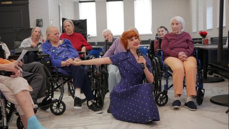 Singer in a vintage blue polka-dot dress kneeling and holding a microphone while reaching to hold hands with elderly residents in wheelchairs in a bright senior living activity room