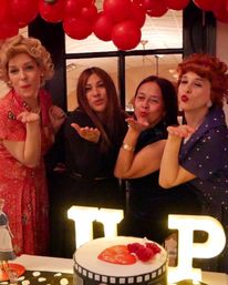 Four people at an indoor celebration blowing kisses under a red balloon garland, wearing vintage-style outfits, with illuminated marquee letters and a decorated cake on the table.