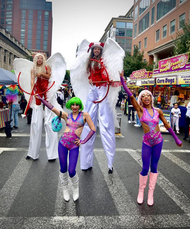 Street performers at a downtown street festival — two stilt-walkers in giant white angel wings and red costumes and two vibrant dancers in neon purple leotards and colorful wigs posing on a city block in front of food vendor booths.