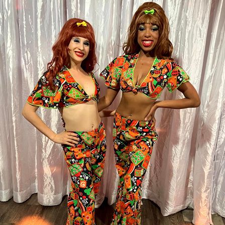 Two performers wearing matching vibrant 1970s-style crop tops and bell-bottoms with orange-green retro print, big wigs with bows, smiling against a pink curtain backdrop.