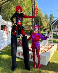 Two colorful jester performers — one on stilts in a black-and-red costume and one in a purple-pink jester outfit with striped tights — posing by a ball pit and inflatable at an outdoor neighborhood family festival