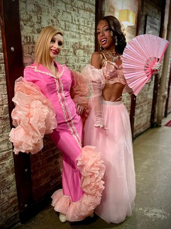 Two performers in pink vintage-inspired costumes pose against a distressed brick wall backstage — one in a ruffled satin suit with dramatic puff sleeves and flared cuffs, the other in a lace bralette, tulle skirt, sheer gloves and holding a glittery pink fan.