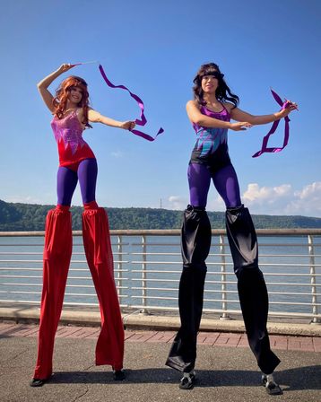 Two stilt performers in vibrant purple and red costumes twirling long purple ribbons on a sunny riverside promenade, metal railing, calm river and tree-covered hills in the background.