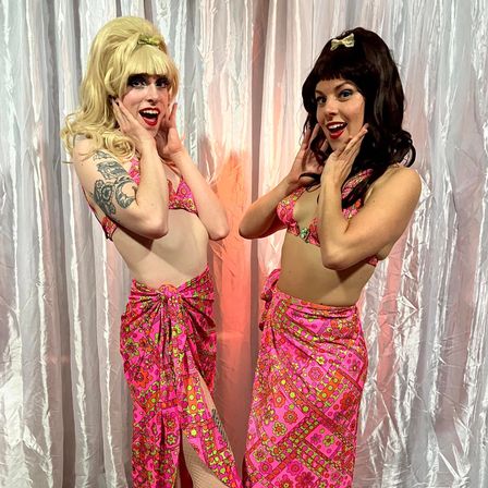 Two performers in matching pink 1960s-style floral bikinis and sarongs, blonde and brunette wigs, striking playful surprised poses in front of a shimmery stage curtain