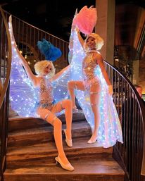 Two showgirl performers in sequined costumes and blonde wigs posing on wooden stairs inside a cabaret-style venue, wearing illuminated LED wings and bright blue and pink feather fans — nightclub entertainment photo.