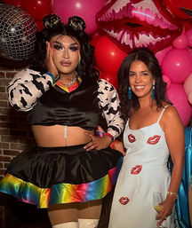 Drag performer in black crop top and rainbow-trimmed skirt with dramatic makeup poses next to a smiling woman in a white dress with red lip appliqués holding a wine glass, with pink lip-shaped balloons and a disco-ball backdrop at a lively party.
