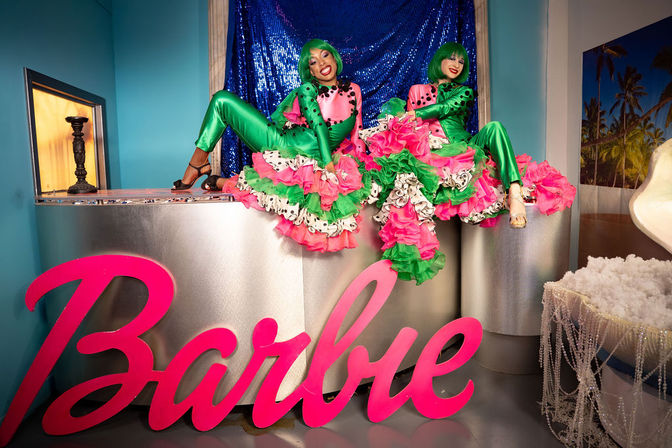 Two performers in green wigs and shiny green bodysuits with pink-and-green ruffled skirts pose on a silver counter in front of a blue sequin backdrop above a large pink "Barbie" sign in a colorful indoor photo set.