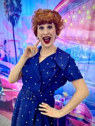 Excited redhead in a vintage 1950s navy polka-dot dress with pearl necklace and bright red lipstick, posing with hand on her cheek against a colorful neon retro diner backdrop with palm trees.