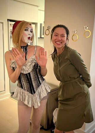 Playful apartment scene: two friends in a modern kitchen — one in silver ruffled costume with white mime makeup and hands raised, the other smiling in an olive-green dress by a wall with gold ring decals.