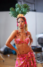 Polynesian-inspired performer in a pink floral bikini and sarong with a large leafy flower headdress, striking a confident pose in an indoor studio
