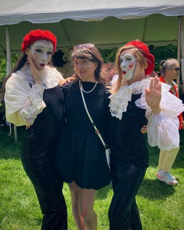 Smiling woman in a black dress and pearl necklace poses between two mimes in red berets, white face paint and ruffled collars at an outdoor park festival tent