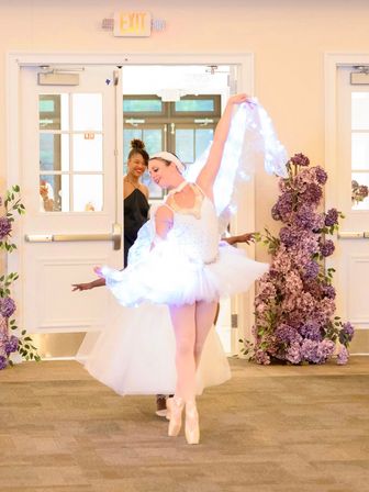 Ballerina in a white tutu and LED-lit veil dancing en pointe through open double doors into an indoor reception, framed by tall purple floral arrangements as a smiling guest watches.