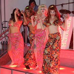 Four costumed performers in vibrant 1960s‑style psychedelic bikini tops and long sarongs dancing on a small illuminated indoor stage with red lighting and marquee bulbs.