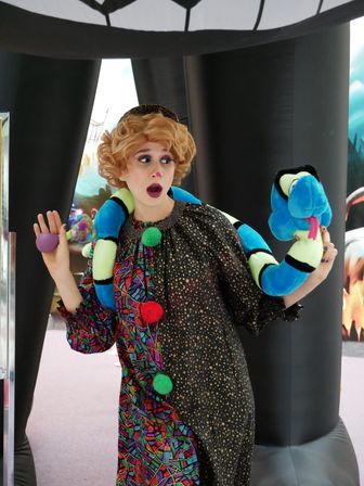 Costumed performer with theatrical makeup and surprised expression, wearing a colorful patterned dress and starry cape, holding a bright blue plush snake draped around their shoulders at an indoor children’s event