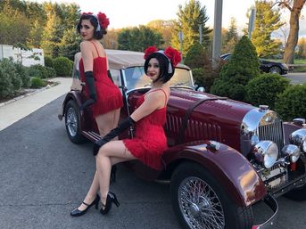 Two performers in red 1920s-style flapper dresses and black gloves posing on a maroon vintage convertible parked on a suburban street at golden hour, surrounded by trees and shrubs.