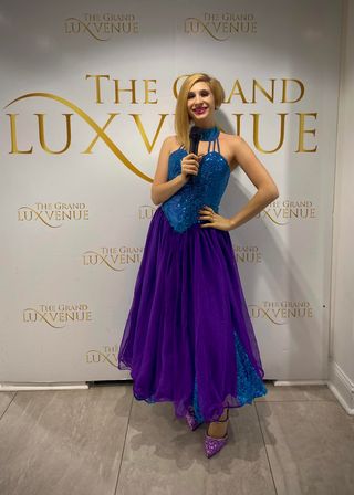 Smiling person in a sparkly blue halter top and flowing purple skirt holding a microphone, posing in front of a gold-lettered event photo backdrop at an indoor venue with glittery heels visible.