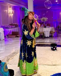 Wizard-costumed entertainer blowing a giant soap bubble in a purple-lit banquet hall with round tables, gold chairs and event decor
