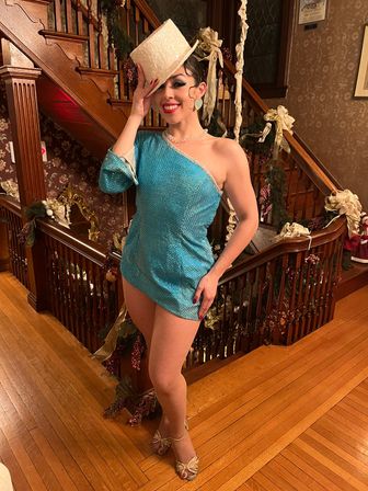 Performer in turquoise sequined one-shoulder dance costume, fishnet tights and white top hat posing on a hardwood floor by a decorated Victorian staircase with holiday garlands and gold bows