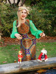 Person in a colorful vintage patterned dress with green sheer sleeves and decorative hat, smiling while holding two marionette puppets (jester and crowned king) above a stone pond edge in a leafy garden.