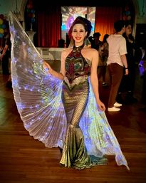 Smiling performer in a sequined mermaid gown and glowing LED wings posing on a wooden dance floor at an indoor party, with colorful balloons and a stage with red curtains in the background.