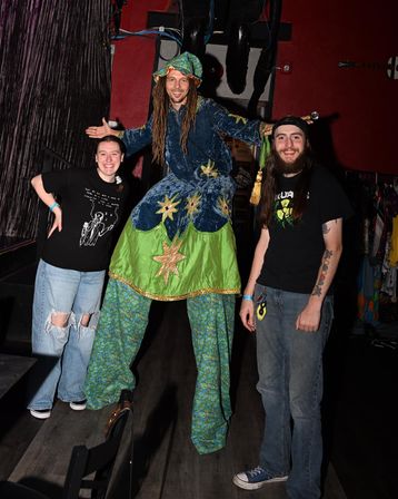 Stilt performer in a blue-and-green starry wizard costume and floppy hat with long dreadlocks poses with two smiling attendees in jeans inside a dimly lit music venue.