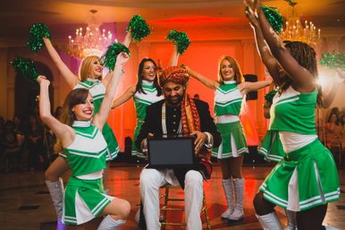 Groom wearing an ornate turban seated in a ballroom opening a gift box while cheerleaders in green uniforms dance around him with pom-poms under crystal chandeliers.