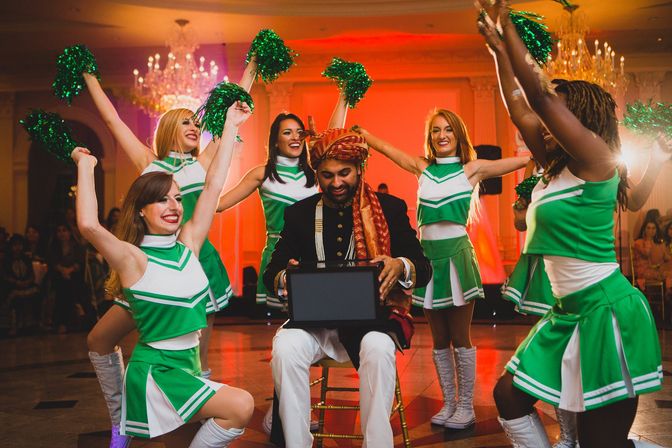 Groom wearing an ornate turban seated in a ballroom opening a gift box while cheerleaders in green uniforms dance around him with pom-poms under crystal chandeliers.