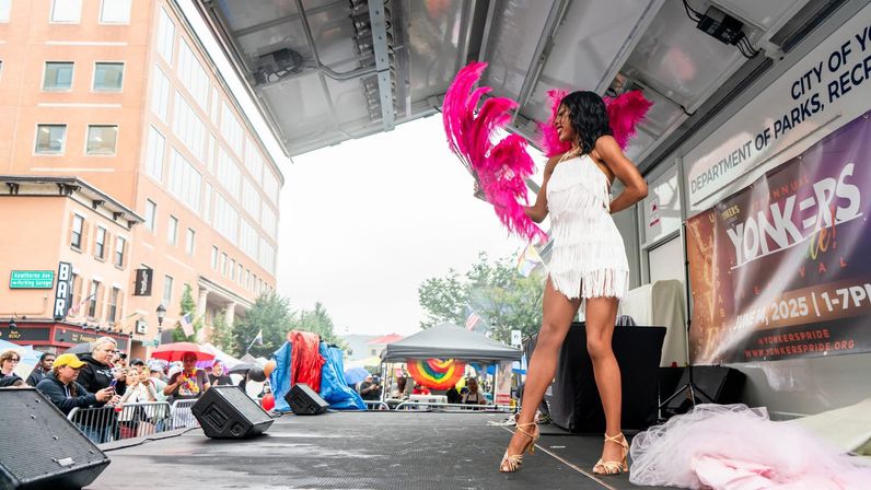 Drag performer in white fringe dress and bright pink feather fans on an outdoor stage entertaining a cheering crowd at Yonkers Pride in downtown Yonkers.