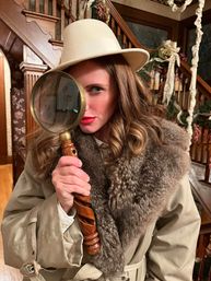 Woman in a beige hat and fur-collared trench coat holding an antique magnifying glass to one eye in a wood-paneled Victorian-style foyer with an ornate staircase