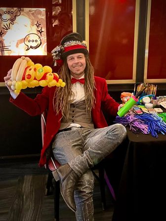 Balloon artist entertainer at an indoor party — smiling performer in a red velvet jacket and decorated top hat holds a yellow balloon crown and green pump beside a table of balloon-twisting supplies.