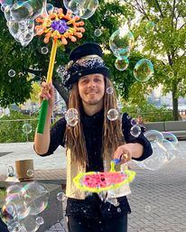 Smiling street performer in a sparkly vest and top hat creating giant iridescent soap bubbles with colorful wands at an urban waterfront park promenade