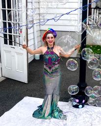 Smiling outdoor bubble performer in a shimmering mermaid-style gown and rainbow headpiece creating giant iridescent soap bubbles in front of a white clapboard building.