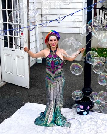 Smiling outdoor bubble performer in a shimmering mermaid-style gown and rainbow headpiece creating giant iridescent soap bubbles in front of a white clapboard building.