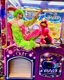 Performer in a green wig and pink-green clown costume posing on a purple arcade prize crane machine with a bright pink puppet bird, colorful lights and prize signage in the background