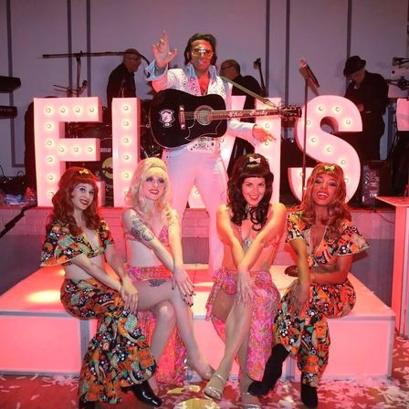Elvis tribute performer in white jumpsuit with guitar standing behind four retro dancers in colorful 1960s‑style costumes, posing on a lit stage with large illuminated marquee letters spelling ELVIS