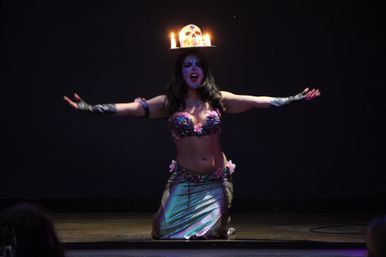 Theatrical performer kneeling on a dark stage in a shimmering belly-dance costume with face paint and outstretched arms, wearing a candlelit skull headpiece.