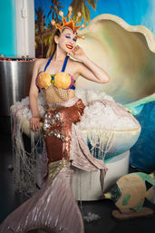 Person in a mermaid costume with seashell bra, coral crown and sequined pink tail seated inside a giant clam prop with pearl decorations against a tropical beach backdrop.