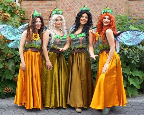 Group portrait of four smiling fairies in green leaf bodices, iridescent blue wings and long golden-orange skirts posing outdoors in a garden in front of a brick wall