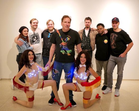 Group photo in an indoor art gallery: eight casually dressed adults standing behind two kneeling showgirl performers wearing light-up white-and-red fringed costumes and red heels, posing against a plain white wall.