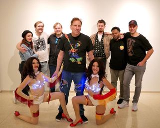 Group photo in an indoor art gallery: eight casually dressed adults standing behind two kneeling showgirl performers wearing light-up white-and-red fringed costumes and red heels, posing against a plain white wall.