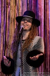 Smiling singer with long dreadlocks and a wide-brimmed hat, wearing a striped jacket with fur cuffs, performing into a microphone against shimmering purple stage curtains.
