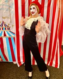 Mime performer in a red beret and white face paint poses with hands up in front of a red-and-white striped carnival backdrop, wearing ruffled sheer sleeves and a black sequined jumpsuit.