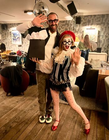 Two people posing in a cozy indoor lounge under a disco ball at a costume party — one casually dressed holding a gift bag, the other in a mime-inspired red beret, striped corset, heart-print tights and red glitter shoes.